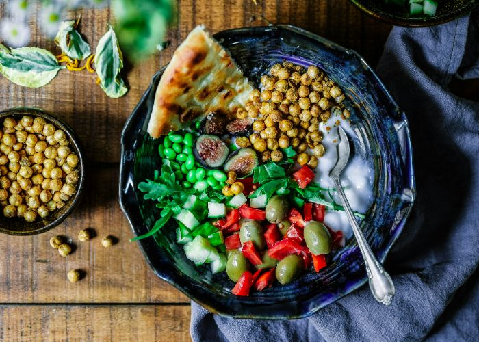 A bowl of different vegetables, including olives, edamame, chickpeas, chilis, cilantro, pita bread, and sauces.